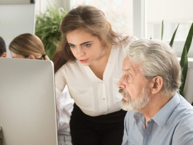 Female executive teaching senior colleague explaining computer work, corporate teacher helping focused aged manager with online task, young woman boss giving instructions to old employee at workplace